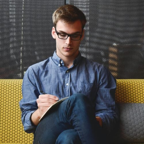 entrepreneur-startup-start-up-man-39866-39866 A young man in glasses writes in a notebook while sitting on a stylish couch indoors.
